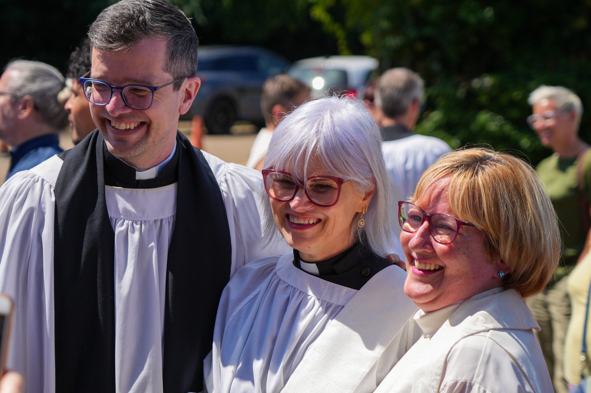 A male and two female clergy in robes smiling outside on a sunny day