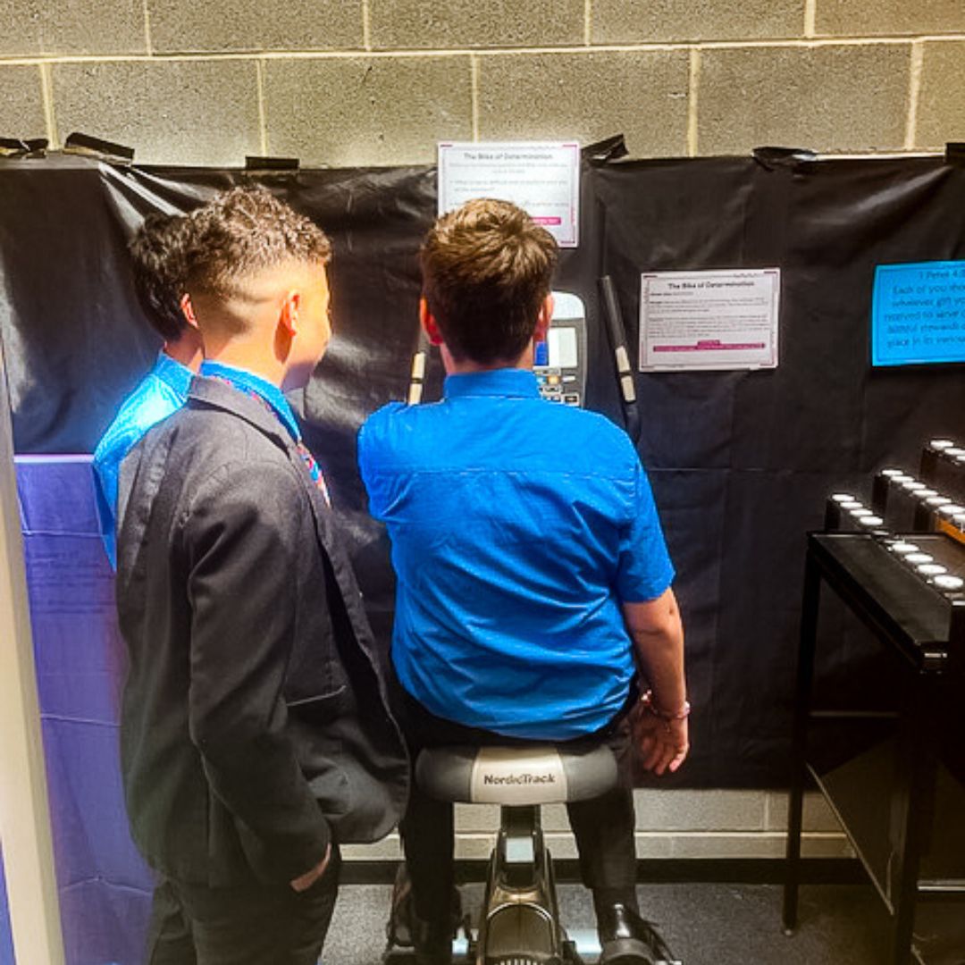 A group of school boys stood around an exercise bike with one sat on