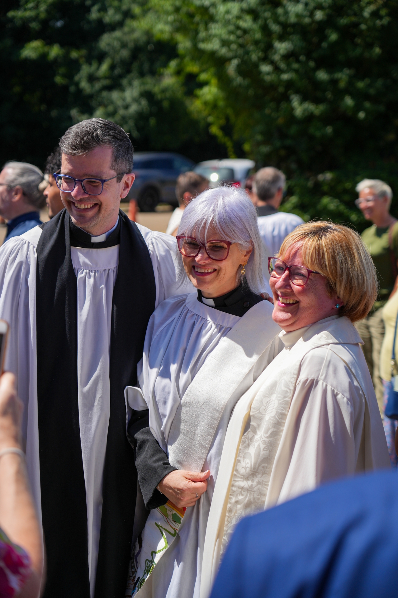 A male and two female clergy robed smiling outside on a sunny day.