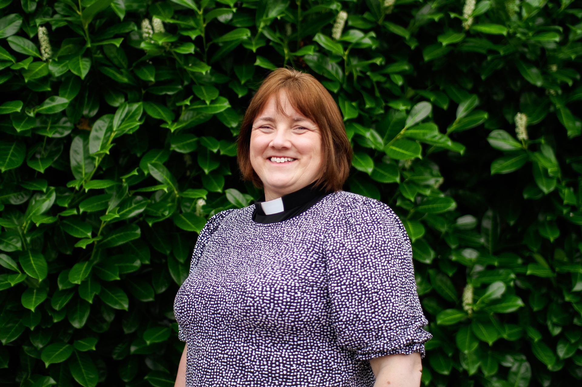 Catharine Mabuza wearing a patterned clerical shirt stood in front of a hedge