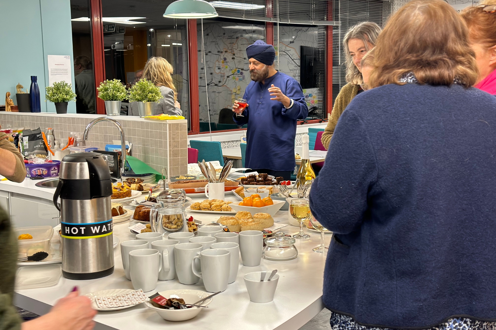 Snack foods on a table, among cups. Man with a beard and blue headdress in conversation with a woman.